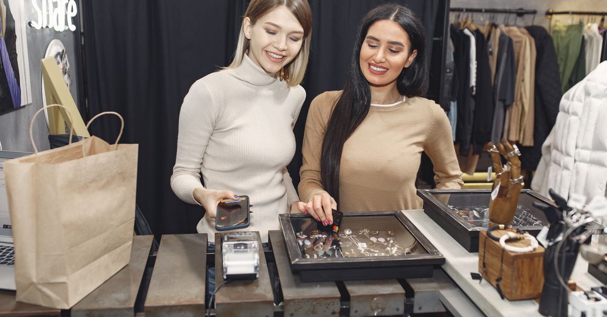 Cheerful casually dressed women paying with smartphone and credit card for purchases while standing near contactless terminal at counter desk